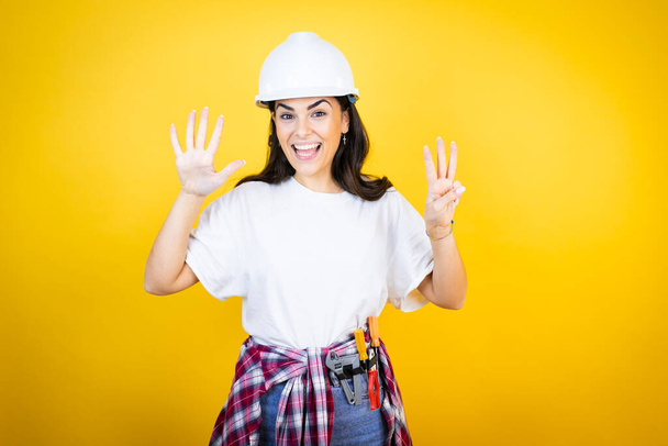 Mladá běloška žena na sobě hardhat a stavitel oblečení přes izolované žluté pozadí ukazuje a ukazuje nahoru s prsty číslo osm a zároveň se usmívá sebevědomě a šťastně - Fotografie, Obrázek