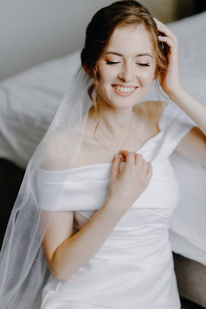 Mattina nuziale della sposa. Squisita sposa dai capelli castani in un abito bianco sorridente alla macchina fotografica. Foto di alta qualità - Foto, immagini