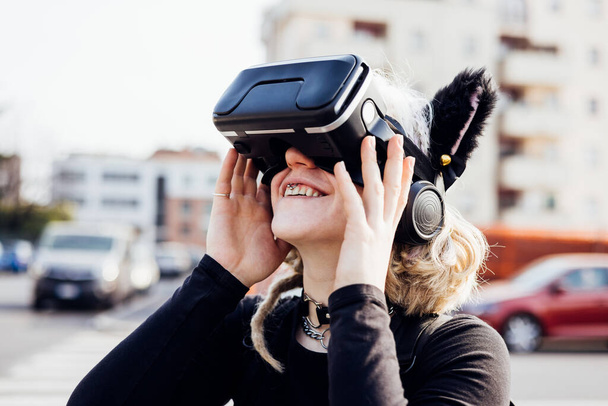 Mujer joven inconformista sonriendo al aire libre usando auriculares de realidad aumentada viendo video disfrutando de la tecnología metaverse 5g - Foto, imagen