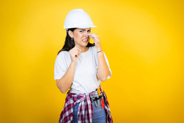 Young caucasian woman wearing hardhat and builder clothes over isolated yellow background smelling something stinky and disgusting, intolerable smell, holding breath with fingers on nose - Foto, Imagem
