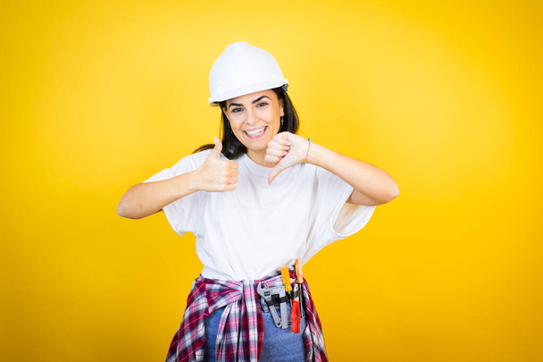 Jonge blanke vrouw draagt hardhoed en bouwer kleding over geïsoleerde gele achtergrond Doen duimen op en neer, onenigheid en overeenstemming uitdrukking. Gekkenhuis - Foto, afbeelding
