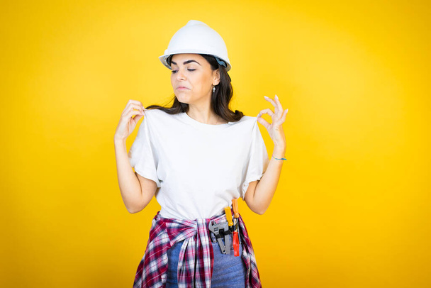 Young caucasian woman wearing hardhat and builder clothes over isolated yellow background holding her t-shirt with a successful expression - Photo, Image