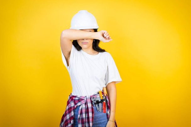 Kafkas şapkalı genç bir kadın izole edilmiş sarı arka planda inşaatçı kıyafetleri giyiyor. Gözleri kollarıyla neşeli ve komik gülümsüyor. Kör kavram. - Fotoğraf, Görsel