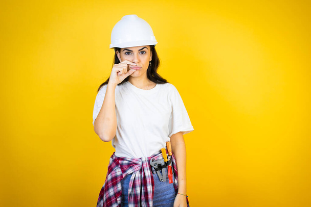 Young caucasian woman wearing hardhat and builder clothes over isolated yellow background mouth and lips shut as zip with fingers. Secret and silent, taboo talking - 写真・画像