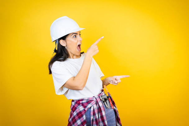 Jeune femme caucasienne portant un casque dur et des vêtements de constructeur sur fond jaune isolé surpris et pointant ses doigts côté - Photo, image