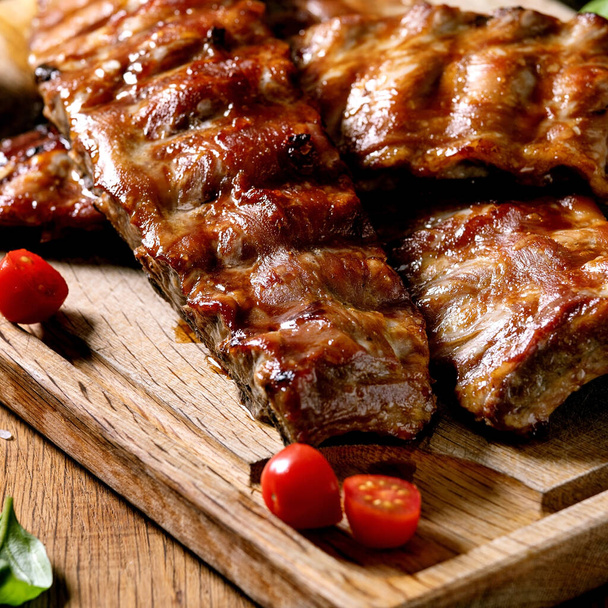 Grilled pork bbq ribs served with cherry tomatoes, basil and barbeque sauce on wooden cutting board on wood board. Close up - Φωτογραφία, εικόνα