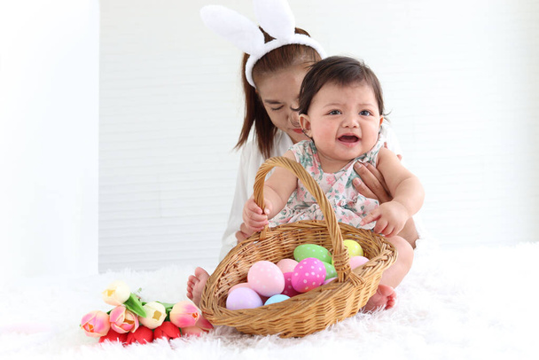 Happy smiling adorable six months baby girl playing with wicker basket of colorful Easter eggs in embrace of mother arms, mom with rabbit ears headband holding her sweet little daughter kid. - Photo, Image