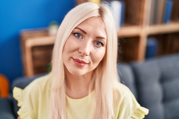 Mladá blondýna žena usměvavá sebevědomě sedí na pohovce doma - Fotografie, Obrázek
