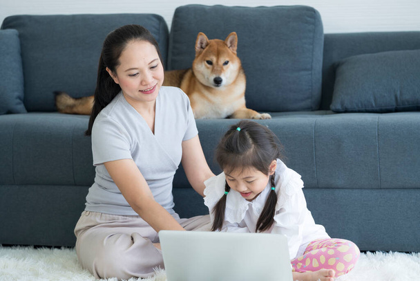 Mother and daughter in the living room with a Shiba Inu sitting on the sofa. Asian mom and little girl look at laptop and shiba dog stay on sofa - Foto, Imagen