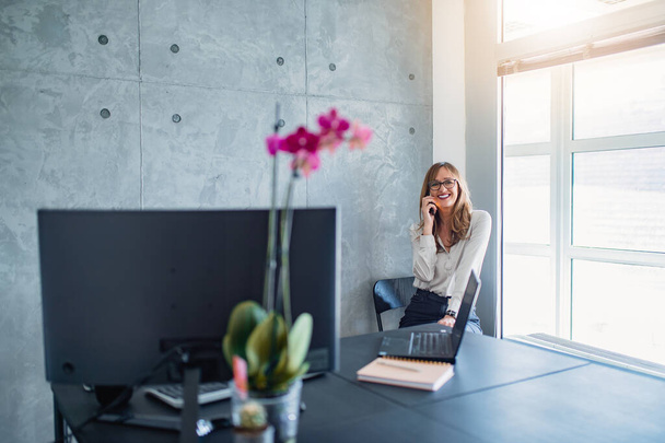 Caucasian middle-aged smiling woman with glasses,sitting on a chair, beside window in  the corner of office,making a phone call.  Vase of pink flower on work desk is perfect detail in grey office.  - Photo, Image