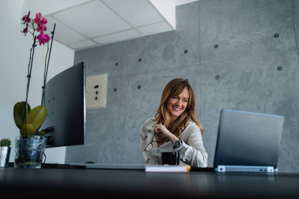 A senior woman has an online meeting and video call on the laptop. White ceiling, gray wall, a vase of flowers, desktop, black cup of some beverage and notebook in office. She smiles and holds eyeglasses in her left hand. - Fotografie, Obrázek