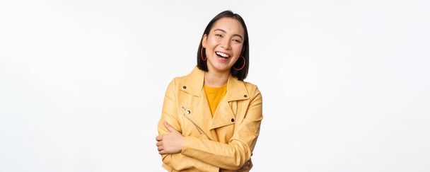 Afbeelding van mooi modern aziatisch meisje lachend, lachend en kijkend gelukkig op camera, staand in geel jasje tegen witte achtergrond - Foto, afbeelding