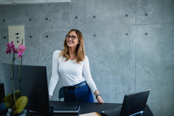 A pretty woman, casual dressed, with modern eyeglasses is standing at her desk, looking at the camera and posing for a photo. The modern interior is part of corporations' offices.  - Foto, afbeelding