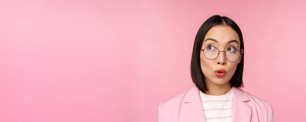 Entusiástica vendedora, asiática empresária de óculos, olhando intrigada no canto superior esquerdo logotipo, anúncio da empresa, de pé sobre fundo rosa - Foto, Imagem
