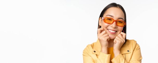 Close up portrait of trendy asian woman in sunglasses, touching her face, looking romantic, smiling at camera, standing over white background - Photo, image