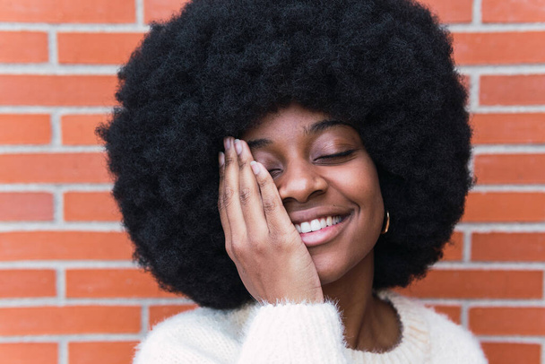 Close up of lovely african american woman with white perfect smile, long afro hair and white jumper covering eye against brick wall. Beautiful afro woman with eyes closed. Beauty concept - Foto, imagen