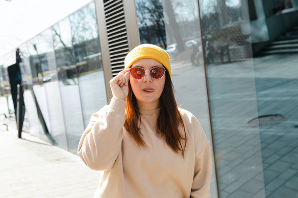 Szczęśliwa młoda, radosna kobieta pokazująca język mrugający na miejskiej ulicy. Portret pięknej kobiety w żółtym kapeluszu i różowych okularach słonecznych uśmiechniętych na zewnątrz. Koncepcja miejskiego stylu życia. Podróżnik - Zdjęcie, obraz