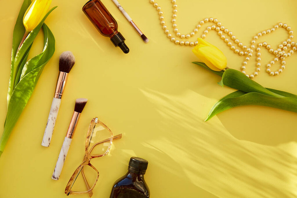Fashionable pastel feminine flat lay: pearls, makeup brushes, glasses, oil, serum dropper. Yellow tulips with trendy shadows. Beauty card concept. Copy space. - Photo, Image