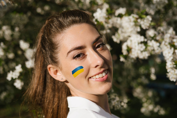 Retrato ao ar livre de mulher ucraniana com bandeira ucraniana azul e amarela em sua bochecha na natureza flor de cereja. Símbolo de liberdade e independência da Ucrânia. Parem a guerra. Esperança e Fé - Foto, Imagem