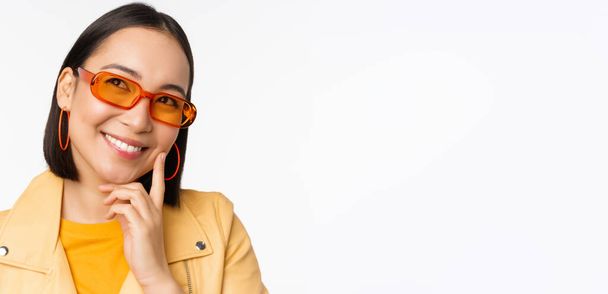 Крупный план портрета азиатской женщины, думающей, одетой в солнечные очки и улыбающейся, смотрящей вдумчиво, стоящей на белом фоне студии - Фото, изображение
