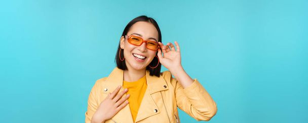 Close up retrato de mulher asiática elegante em óculos de sol, rindo e sorrindo, olhando feliz, posando em roupas da moda sobre fundo azul - Foto, Imagem