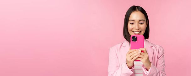 スーツ姿でアジア系企業の女性を笑顔にしたり、スマートフォンアプリを見たり、携帯電話アプリを使ってピンクの背景に立っている姿 - 写真・画像