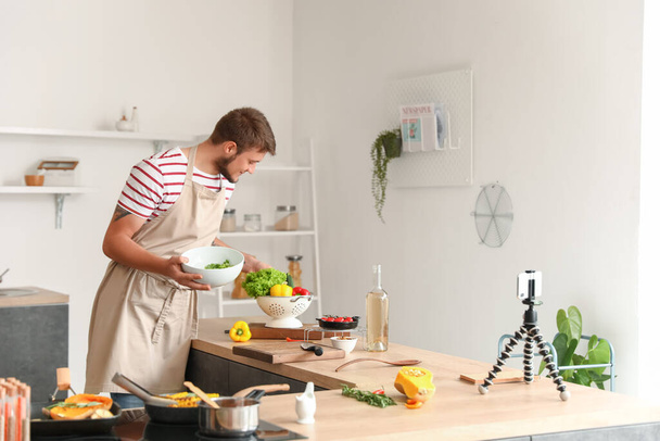 Junger Mann macht Salat, während er Kochvideo-Anleitung in der Küche aufzeichnet - Foto, Bild