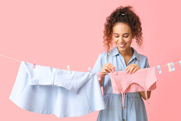 Young African-American woman hanging clean top with clothespin on pink background - Foto, afbeelding