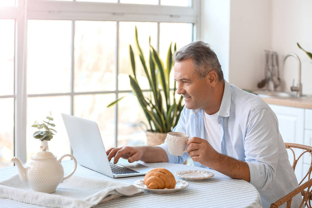 Hombre mayor con taza de té usando portátil en la mesa en la cocina - Foto, imagen
