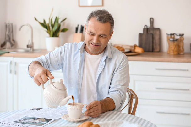 Старший человек наливает чай в чашку за столом на кухне - Фото, изображение