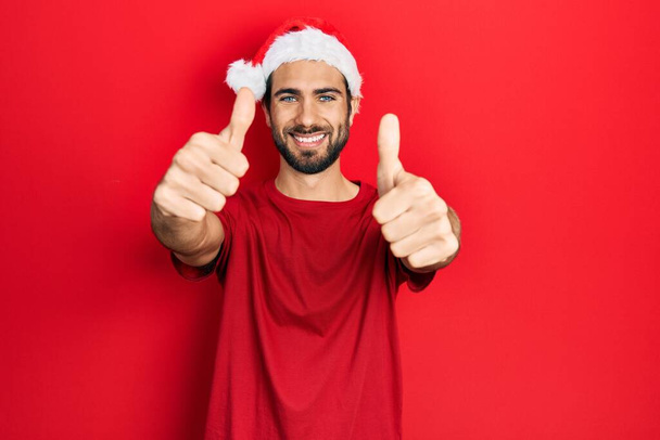 Jeune homme hispanique portant le chapeau de Noël approuvant faire un geste positif avec la main, pouces levés souriant et heureux pour le succès. geste gagnant.  - Photo, image
