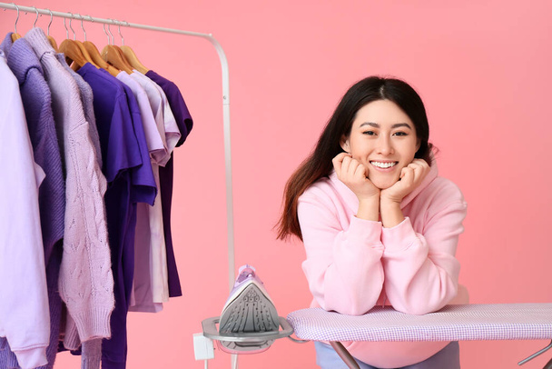 Young Asian woman with rack, clothes and ironing board on pink background - Фото, зображення