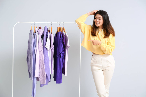 Young Asian woman and rack with clothes in purple shades on light background - Foto, imagen