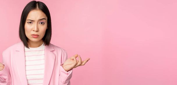 Portrait of young asian business woman, saleswoman shrugging shoulders and looking confused, clueless of smth, standing over pink background - Foto, imagen
