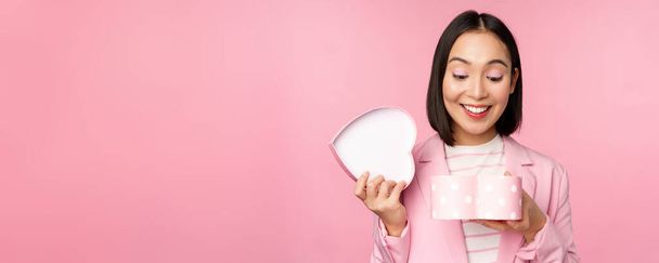 Счастливая симпатичная девушка в костюме, открывает коробочку в форме сердца с романтическим подарком на праздник белого дня, стоя в костюме на розовом фоне - Фото, изображение