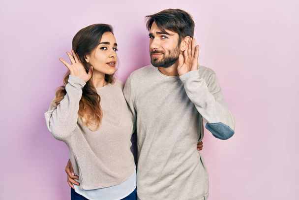 Jovem casal hispânico vestindo roupas casuais sorrindo com a mão sobre o ouvido ouvindo um rumor ou fofoca. conceito de surdez.  - Foto, Imagem