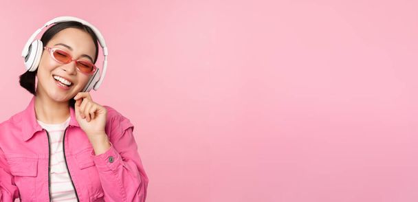 Χορεύοντας κομψό κορίτσι της Ασίας ακούγοντας μουσική στα ακουστικά, θέτοντας σε ροζ φόντο - Φωτογραφία, εικόνα