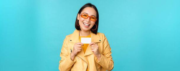 Стильная молодая азиатка в солнечных очках, с кредитной картой и улыбкой, рекомендует банк, бесконтактную оплату или скидки в магазине, стоя на синем фоне - Фото, изображение