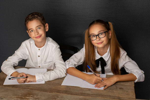 Twee lachende elegante kinderen aan tafel schrijven met pen, op zwarte muur - Foto, afbeelding