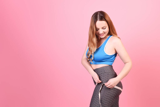 A beautiful body-positive girl on a pink background in a tracksuit measures her waist and hips with a centimeter. Sports and training - Photo, Image