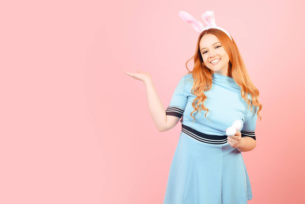 Una chica con el pelo rojo en un vestido azul y orejas de conejo de Pascua sobre un fondo rosa. Una mujer hermosa está apuntando a un producto o texto. - Foto, imagen