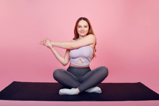 Belle fille positive sur un fond rose dans un survêtement va dans le sport, échauffement des sports de remise en forme, assis sur un tapis de yoga. - Photo, image