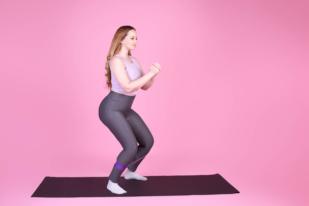 A beautiful body-positive girl on a pink background in a full-length sports suit goes in for sports, elastic bands for training, leg exercises, fitness - Foto, Imagem