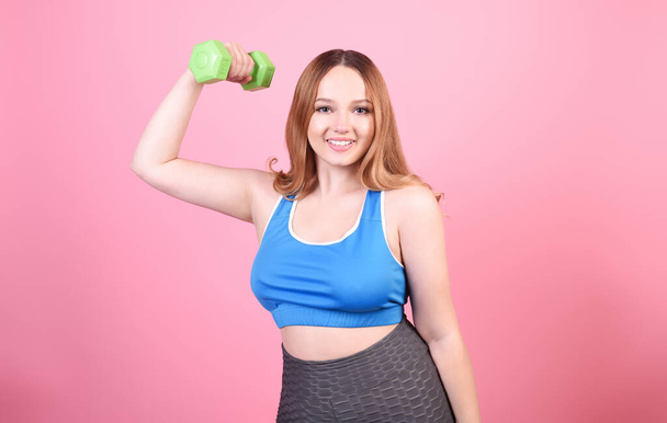 A beautiful body-positive girl on a pink background in a tracksuit lifts weights. Sports, hand training. - Foto, immagini