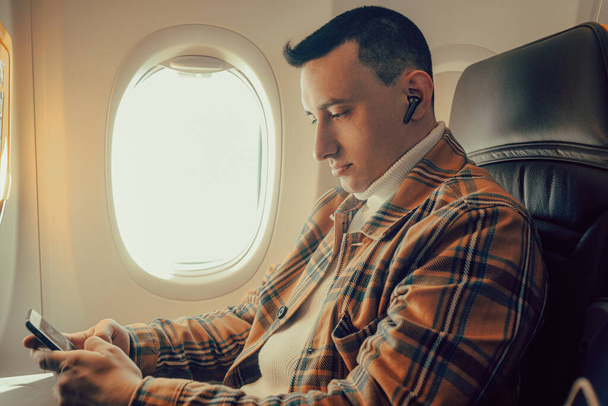 Ein Mann sitzt im Flugzeug am Fenster und blickt in ein Smartphone. - Foto, Bild