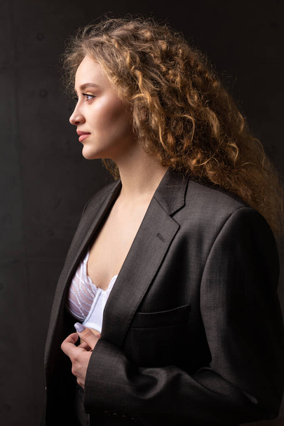 Portrait of a young red-haired curly sexy girl in a jacket with long hair in the studio. Shows off her curvaceous breasts in a bra. - Foto, imagen