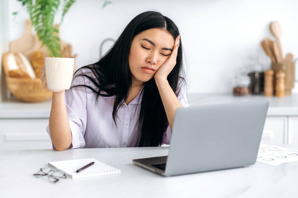Uma menina asiática sonolenta, freelancer ou um estudante, senta-se em casa na cozinha em uma mesa com os olhos fechados na frente de um laptop, segura uma xícara de café na mão, não pode acordar e começar a trabalhar - Foto, Imagem