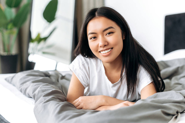Portret van een mooi positief Aziatisch brunette meisje in een wit t-shirt, thuis liggend in gezellig bed, kijkt naar de camera, glimlacht vriendelijk. Ik ben thuis. Relax concept - Foto, afbeelding