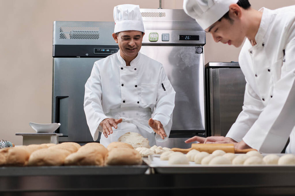Dos cocineros masculinos asiáticos profesionales en uniformes blancos y delantales están amasando masa de pastelería y huevos, preparando pan y alimentos frescos de panadería, horneando en el horno en la cocina de acero inoxidable del restaurante. - Foto, Imagen