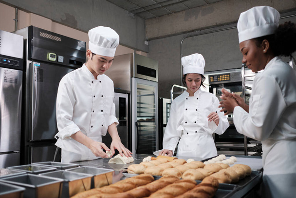 Profesionální gurmánský tým, tři mladí kuchaři v bílých kuchařských uniformách a zástěrách hnětou těsto a vejce, připravují chléb a čerstvé pečivo, pečou v troubě v kuchyni restaurace z nerezové oceli. - Fotografie, Obrázek